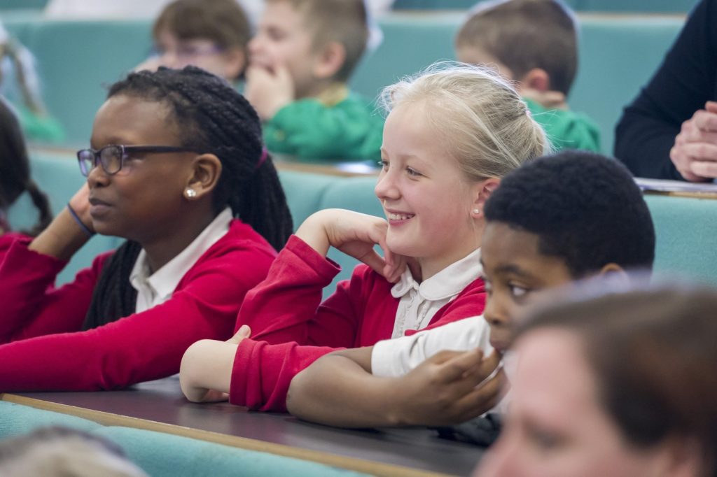 Group of children sat in a lecture theatre smiling and looking to the left.